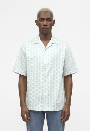 Νέος άνδρας με κοντά ξανθά μαλλιά που φοράει ανοιχτό γκρι κοντομάνικο πουκάμισο με διακριτικό κυκλικό μοτίβο και μπλε τζιν.