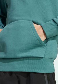 Ljus teal sweatshirt med ribbad nederkant och ärmar; har en framficka med mjuk textur. Materialet verkar vara bomull eller bomullsblandning.