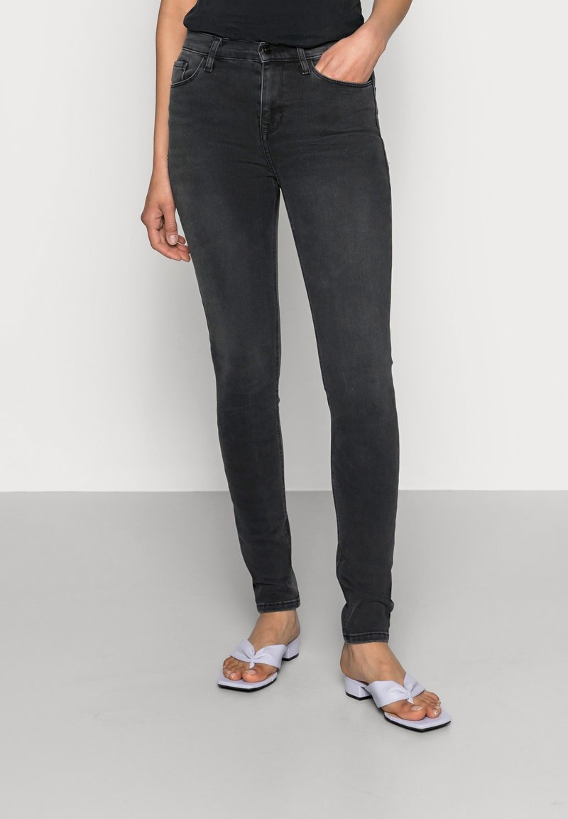 Jean skinny en denim noir avec une texture lisse, taille haute, passants de ceinture et design classique à cinq poches. Associé à des talons à brides en nœud de couleur violet clair.