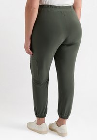 Gröna joggingbyxor med elastisk midja, sidoficka, avsmalnande anklar och ett mjukt, lätt tyg. Bärs med beige sneakers.