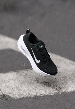 Zapatilla de correr Nike en blanco y negro en el aire sobre asfalto con una marca de flecha blanca, mostrando el lateral y la suela con amortiguación ZoomX.
