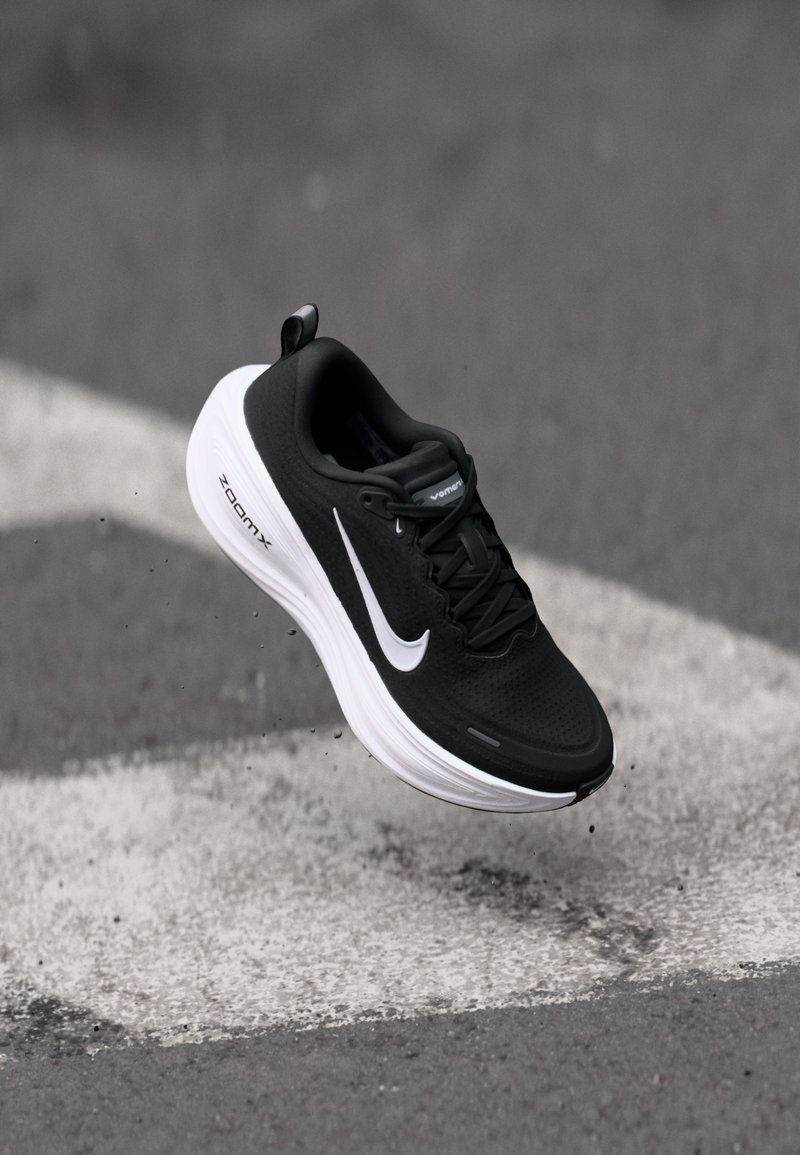 Chaussure de course Nike noire et blanche en l'air au-dessus de l'asphalte, avec un marquage flèche blanche, montrant le côté et la semelle avec amorti ZoomX.