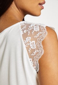 Vit topp med broderad spetsdetalj på axeln, med vågiga kanter och mjuk textur som framhäver en delikat design.