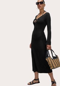 Černé šaty s dlouhým rukávem, výstřihem do V a zavazováním, doplněné tkanou taškou a sandály na ploché podrážce. Hladká textura a střih do poloviny délky.