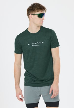 Ung mand iført grøn "Endurance" t-shirt, grå træningsshorts med sorte leggings og refleks sports solbriller, der kigger mod højre.