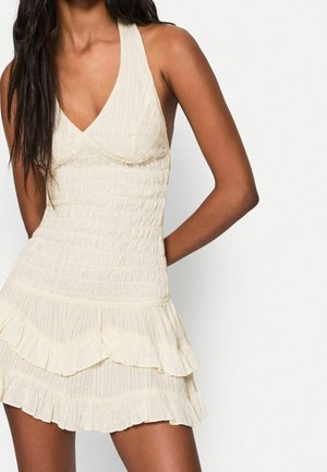 Kremowa mini sukienka bez rękawów z marszczonym gorsetem, plisowaną spódnicą i falbanką na dole, noszona przez kobietę o długich ciemnych włosach.