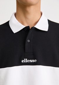 Juodas polo marškinėliai su baltu apykaklė, turintys horizontalią baltą juostą ir išsiuvinėtą baltą "ellesse" logotipą, pagaminti iš medvilnės.