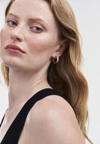 Donna con pelle chiara, lentiggini e capelli castano chiaro ondulati, che indossa un top senza maniche nero e un orecchino a cerchio d'oro con piccole pietre.