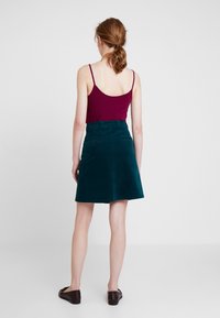 Jupe en velours côtelé teal avec une forme évasée, deux poches arrière avec des boutons, portée avec un débardeur bordeaux, bretelles courtes visibles.