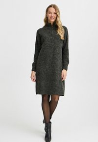 Abito grigio a maglia con colletto alto e zip. Presenta maniche lunghe, una vestibilità comoda e arriva sopra il ginocchio. Indossato con stivali neri alla caviglia.