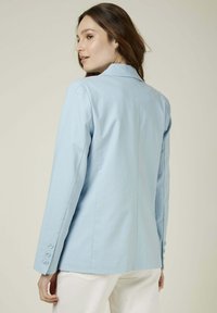 Blazer ajusté bleu clair avec une texture lisse, des épaules structurées, des manches longues et quatre boutons sur chaque poignet, présentant un dos design épuré.