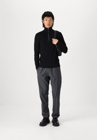 Pull à col roulé noir côtelé avec une fermeture éclair à l'avant, associé à un pantalon à carreaux gris et des chaussures noires à bout rond. Sac noir simple sur l'épaule.