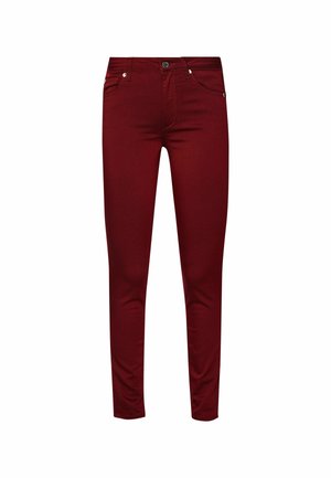 Femme portant un manteau noir, un top noir avec un logo doré, un pantalon slim bordeaux et des mocassins noirs, debout devant un fond clair uni.