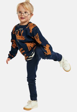 Laivaston sininen collegepaita, jossa ruskean kissan kuvio, yhdistettynä laivastonsinisiin housuihin. Valkoiset tennarit kumipohjalla. Silmälasit päässä, lapsi poseeraa leikkisästi.