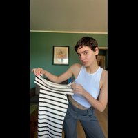 Camicia a righe bianca e nera, tessuto leggero, vestibilità comoda. Modello che indossa un top bianco senza maniche, in piedi in una stanza con pareti verdi.