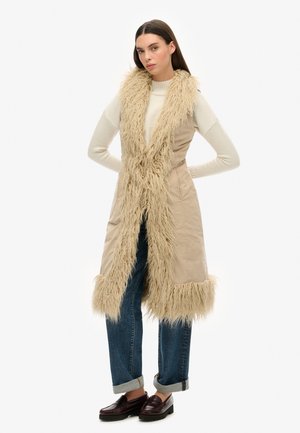 Beige langer Weste mit einem Kragen und Besatz aus Kunstfell, kombiniert mit einem engen Pullover, dunkelblauen Jeans und dunklen Slippern.