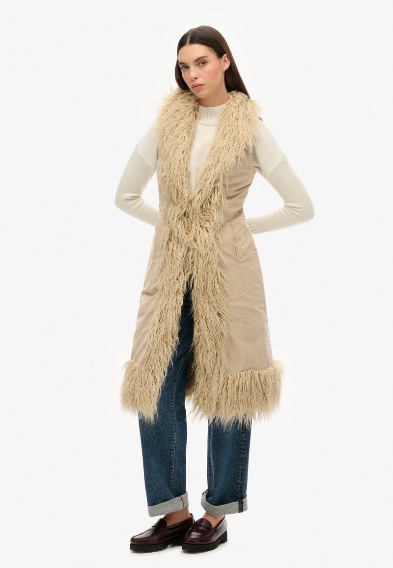 Beige langer Weste mit einem Kragen und Besatz aus Kunstfell, kombiniert mit einem engen Pullover, dunkelblauen Jeans und dunklen Slippern.
