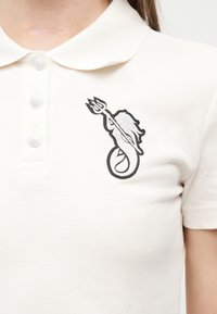 Λευκό πόλο πουκάμισο με τρία κουμπιά και μια μαύρη γοργόνα που κρατά ένα τρίαινα κεντημένη στο αριστερό στήθος.