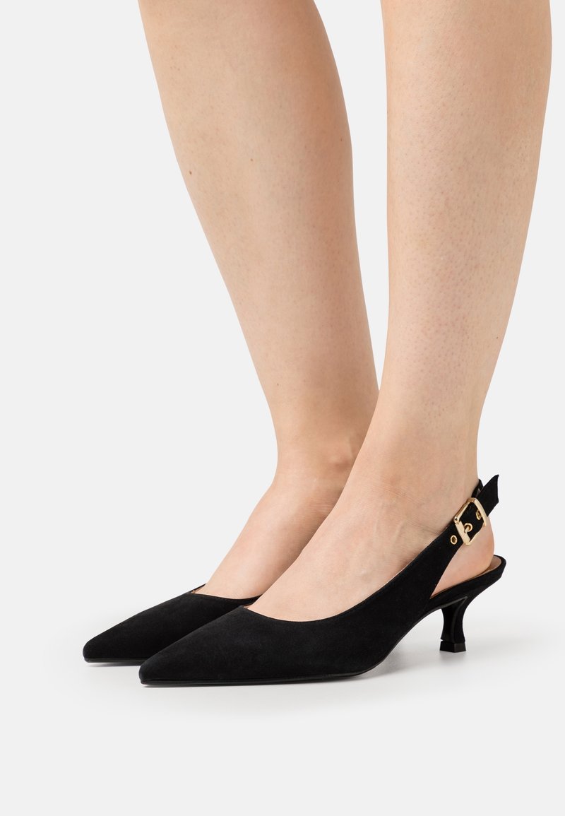 Jambes portant des escarpins slingback en daim noir à bouts pointus, avec petits talons kitten et détails de boucles dorées.