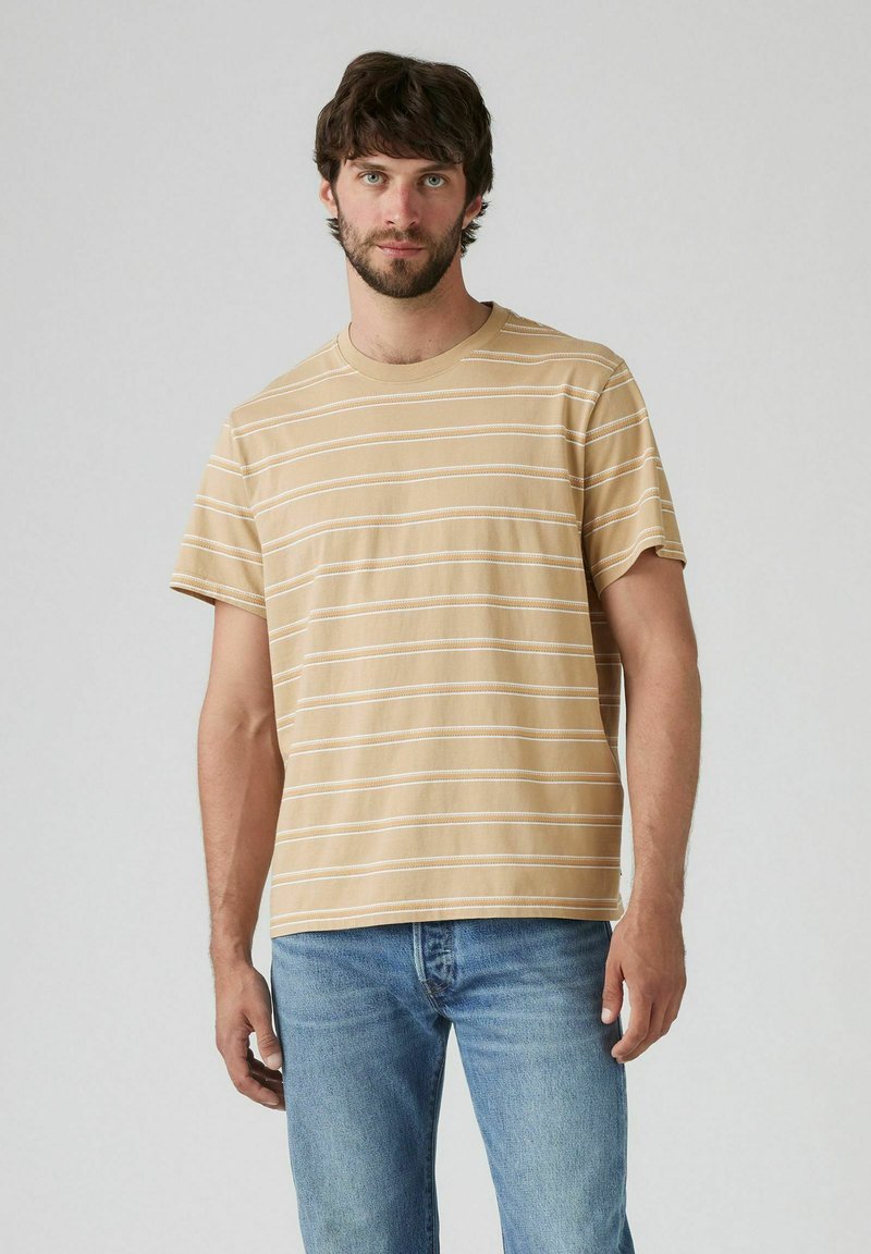 Beżowa bawełniana koszulka z poziomymi białymi paskami, okrągłym dekoltem, krótkimi rękawami i luźnym krojem, noszona z jasnoniebieskimi dżinsami.