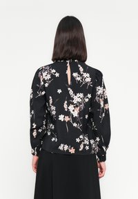 Donna con capelli scuri e lisci indossa una blusa nera a fiori con maniche lunghe e una piccola apertura a chiave sul retro, abbinata a una gonna nera.