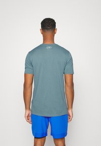 Pánské tričko s krátkým rukávem a kulatým výstřihem v barvě teal. Na zadní straně se nachází umístěné logo a tričko je kombinováno s modrými sportovními šortkami.