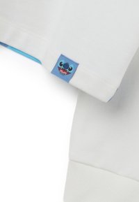 Camiseta de algodón blanca con una etiqueta de tela azul que presenta la cara de un personaje de dibujos animados. La etiqueta está cosida en el dobladillo inferior, contrastando con el tejido.