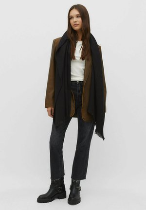 Veste en cuir marron avec des coutures orange, haut blanc, écharpe noire, jeans noirs taille haute et bottines noires à boucles.
