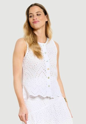 Vrouw draagt een mouwloze witte blouse met oogjes en knopen en een bijpassende rok, kijkt opzij met lang golvend haar.
