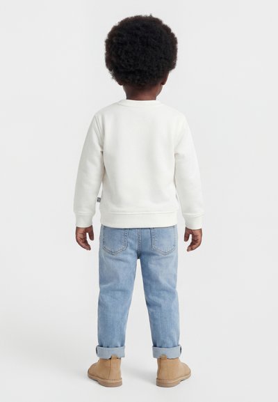 Kind mit lockigem Haar, trägt weißen Pullover, hochgekrempelte hellblaue Jeans und beige Stiefel, steht mit dem Rücken zur Kamera vor weißem Hintergrund.
