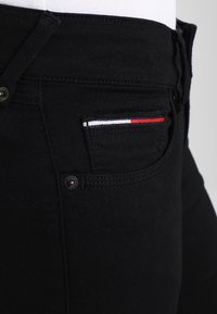 Zblízka na kapsu černých jeansů s malým obdélníkovým textilním detailem v červené, bílé a modré barvě na horním okraji kapsy.