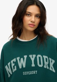 Felpa verde con la scritta "NEW YORK" e "SUPERDRY" in grande, in un grigio chiaro. Collo a costine e vestibilità comoda con una texture morbida.