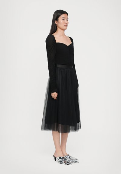 Zwarte top met lange mouwen en geplooide details, gecombineerd met een zwarte tule midi rok. Model draagt zilveren hoge muiltjes met spitse neuzen.