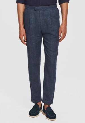 Mężczyzna ubrany w dopasowane ciemnoszare spodnie z podwiniętymi nogawkami i granatowe mokasyny, stojący na jednolitym jasnym tle.