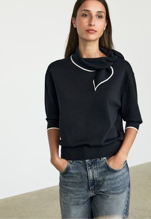 Donna che indossa un maglione nero a maniche corte con rifiniture bianche e dettaglio nodo sul colletto, abbinato a jeans denim azzurro chiaro. Mani nelle tasche.