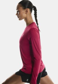 Femeie cu păr împletit, purtând un tricou roșu cu mâneci lungi și pantaloni scurți negri, în poziția de întindere lunge pe un fundal alb.