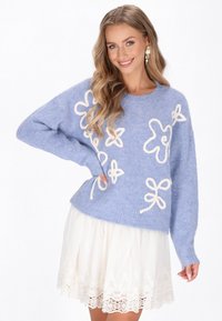 Ljust blått stickat tröja med krämfärgade blommor och rosett-mönster, med rundad halsringning och lös passform, matchad med en vit spetskjol.