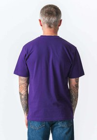 Mand med kort gråt hår og tatoverede arme iført en enkel lilla t-shirt og blå jeans, der vender væk mod en hvid baggrund.