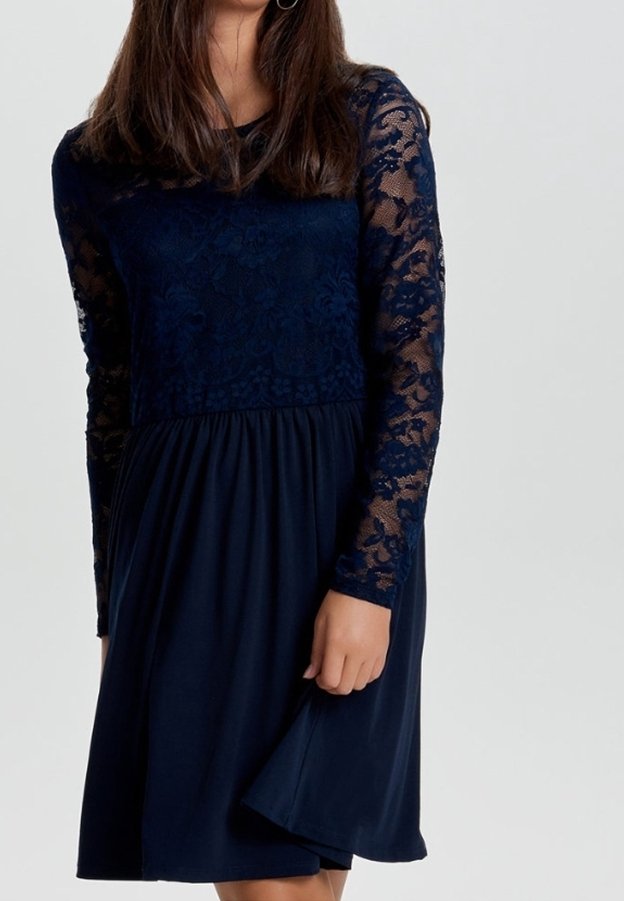 Femme portant une robe bleu marine longueur genou avec manches longues en dentelle et corsage, tenant la jupe d'une main sur un fond uni.