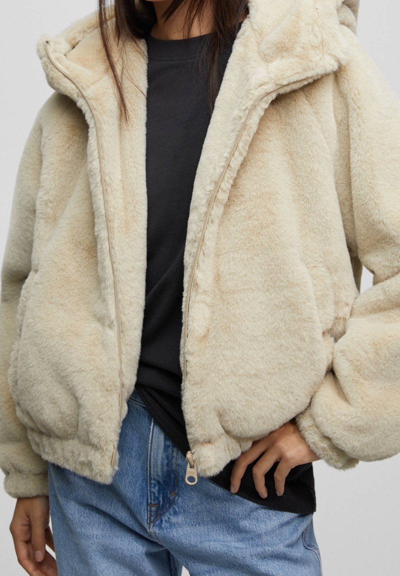 Personne portant une veste beige duveteuse zippée par-dessus une chemise noire et un jean bleu, une main dans la poche de la veste.