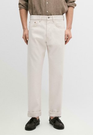 Mężczyzna noszący jasnobeżowe proste spodnie z podwiniętymi mankietami, połączone z brązową koszulą na guziki i czarnymi sznurowanymi butami.