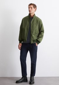 Olive green bomber jacket with a high collar, side pockets, and ribbed cuffs, paired with navy trousers and black ankle boots.