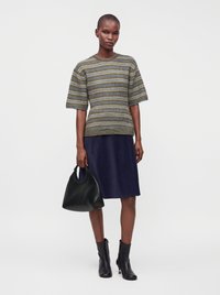 Femme portant un pull à manches courtes rayé, jupe bleu marine longueur genou, bottines noires, tenant un sac à main noir, debout sur fond blanc.