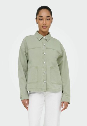 Vrouw met haar naar achter gebonden, draagt een lichtgroene jas met knopen en witte broek, kijkt recht vooruit tegen een effen achtergrond.