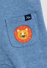 Modrá bavlnená látka má malú zadnú vrecko s výšivkou leva v oranžovej a žltej farbe a tmavomodrou etiketou s nápisom "Jojo."