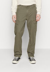 Olijfgroene cargo broek met zijzakken, rechte pasvorm, gemaakt van katoenmix. Draagt met zwart-witte canvas sneakers.