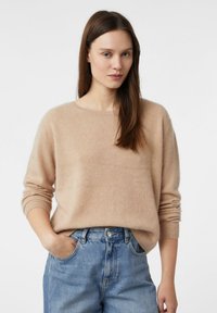 Beżowy sweter z dzianiny o okrągłym dekolcie, z prążkowanymi mankietami i luźnym krojem, zestawiony z wysokim stanem niebieskich dżinsów i rękami w kieszeniach.