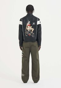 Giacca di pelle nera con accenti bianchi e un grande grafico colorato sul retro, abbinata a pantaloni larghi verde oliva.