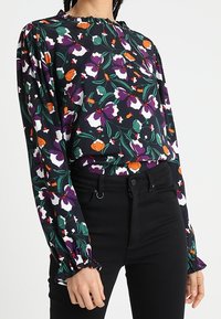 Sort langærmet bluse med et blomstermønster i lilla, orange og hvid. Har en sammensamlet halsudskæring og elastiske manchetter, lavet af letvægtsmateriale.