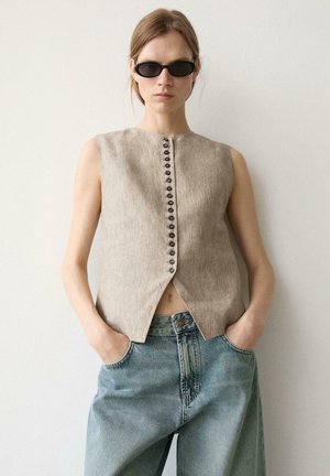 Donna che indossa un gilet beige senza maniche con bottoni, jeans blu larghi a vita alta e occhiali da sole neri ovali, in piedi con le mani in tasca.